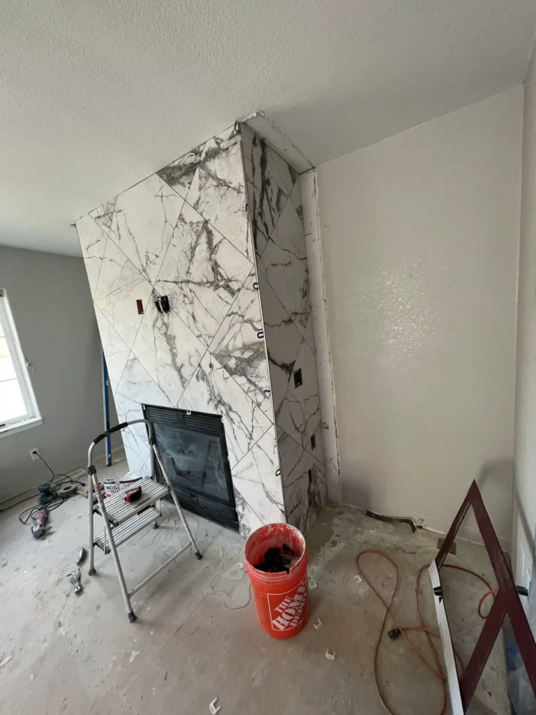 A partially renovated room featuring a fireplace covered with large, white marble tiles with dark veins. A ladder and construction materials are scattered around, indicating ongoing work.