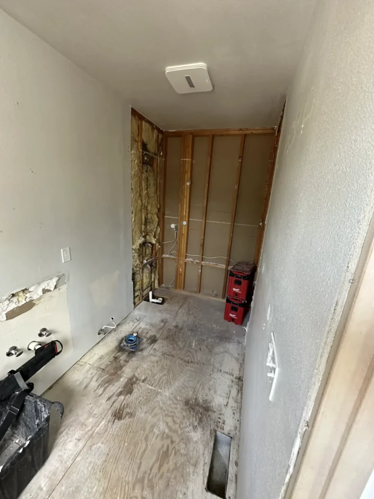 Partially renovated room with exposed studs and insulation, featuring a plywood floor. Plumbing and electrical installations are in progress, with tools and materials on-site.