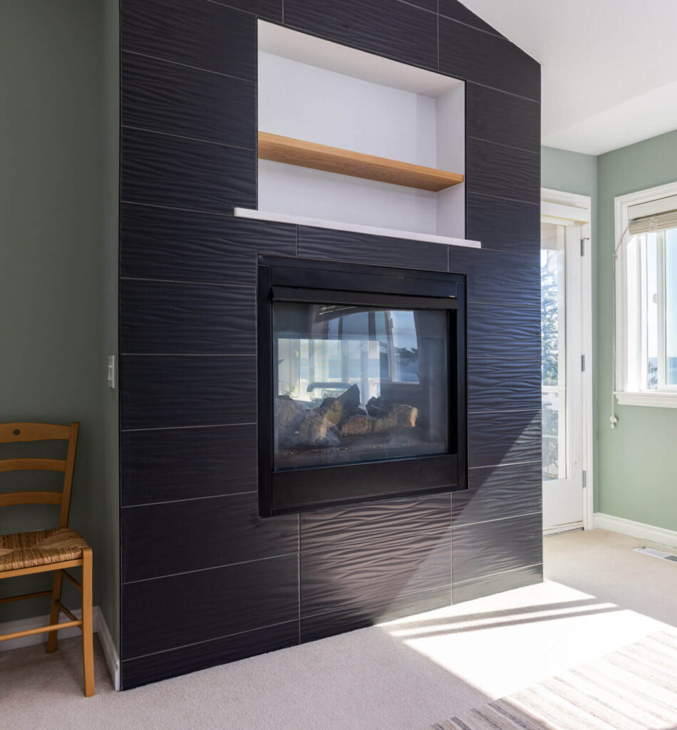 Modern fireplace with a sleek black tile surround in a bright room. The setup includes a built-in shelf above the fireplace and a scenic view through the window.
