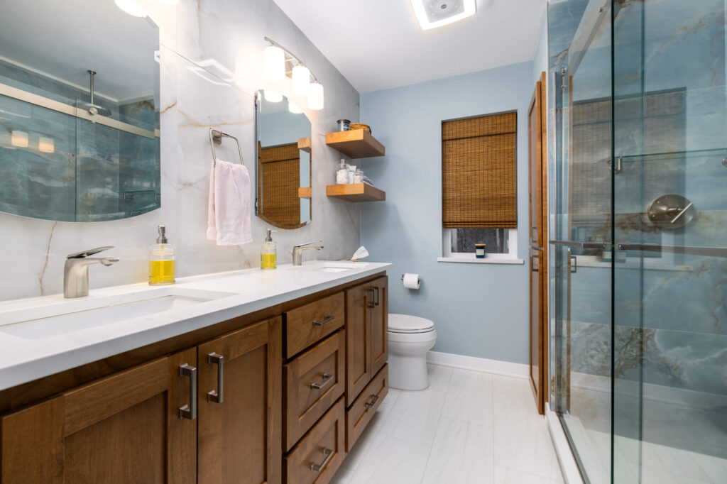Modern bathroom with double sink vanity, marble countertops, and wooden cabinets. Glass-enclosed shower with blue tiles, towel on chrome holder, floating wooden shelves above the toilet, light blue walls, and white tiled floor.