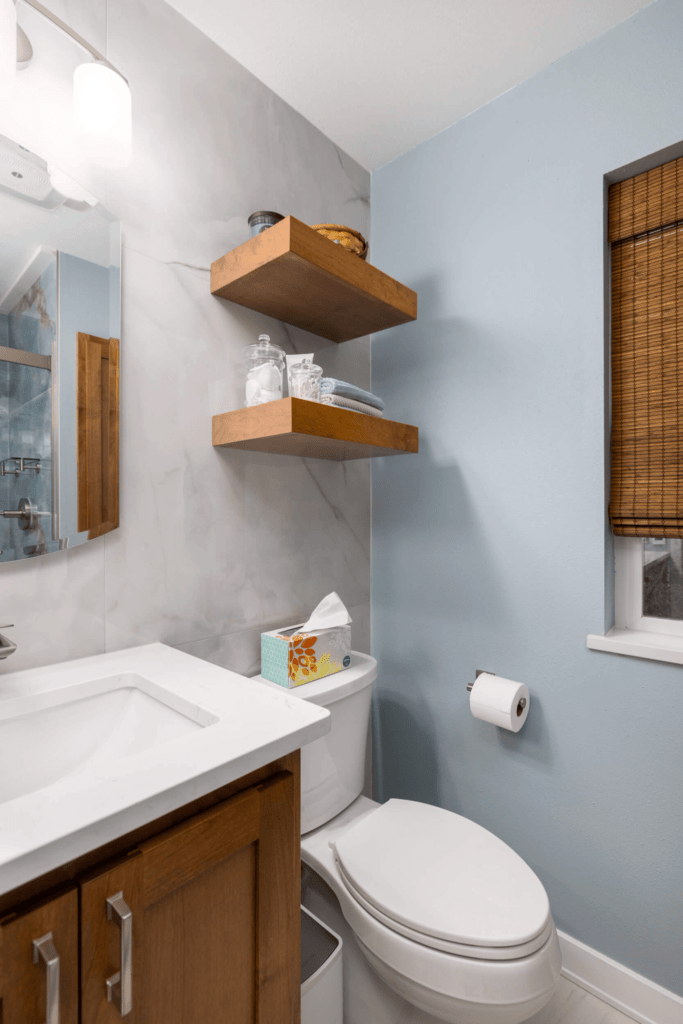 Cozy bathroom interior with light blue walls featuring a white sink, wooden cabinet, and mirror, with two wooden floating shelves above a toilet and a bamboo blind covering a small window.
