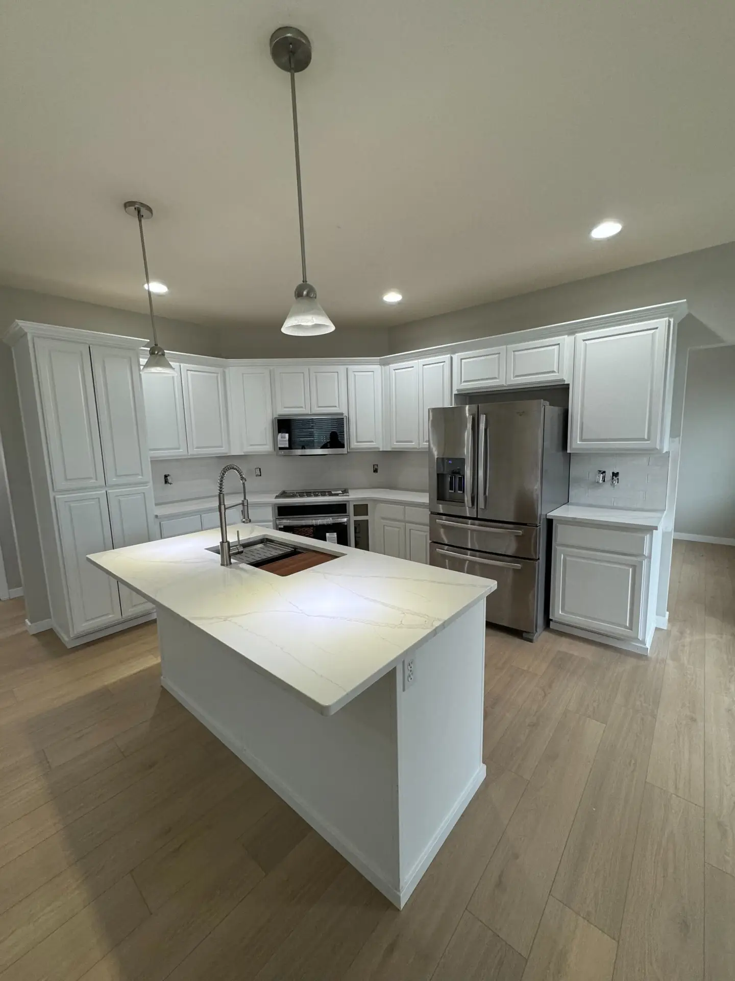 Modern kitchen interior with white cabinetry, stainless steel appliances, an island with a sink, under-cabinet lights, and hanging pendant lights, set against a neutral wall and light wood flooring.