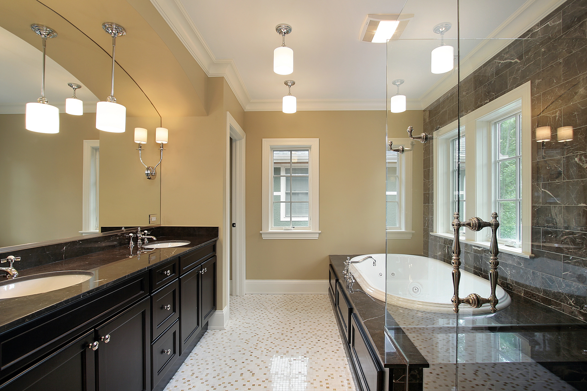 Modern bathroom with double sink vanity, dark cabinets, large mirror, soaking tub by windows, glass shower, and beige walls with white trim. Bright light fixtures hang from the ceiling.