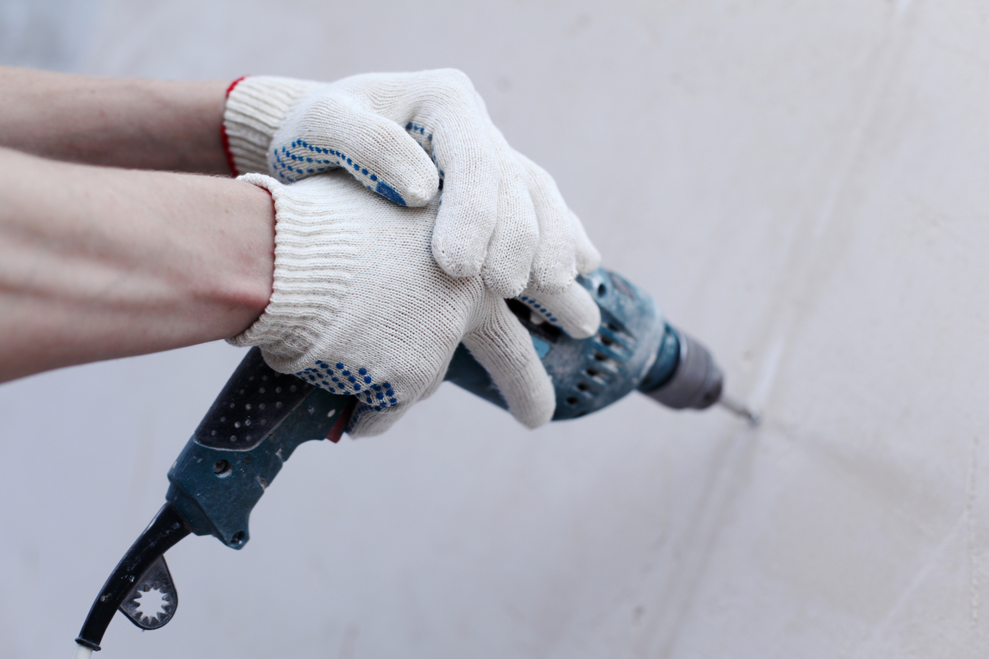 A person wearing white work gloves uses a blue electric drill to make a hole in a white wall.