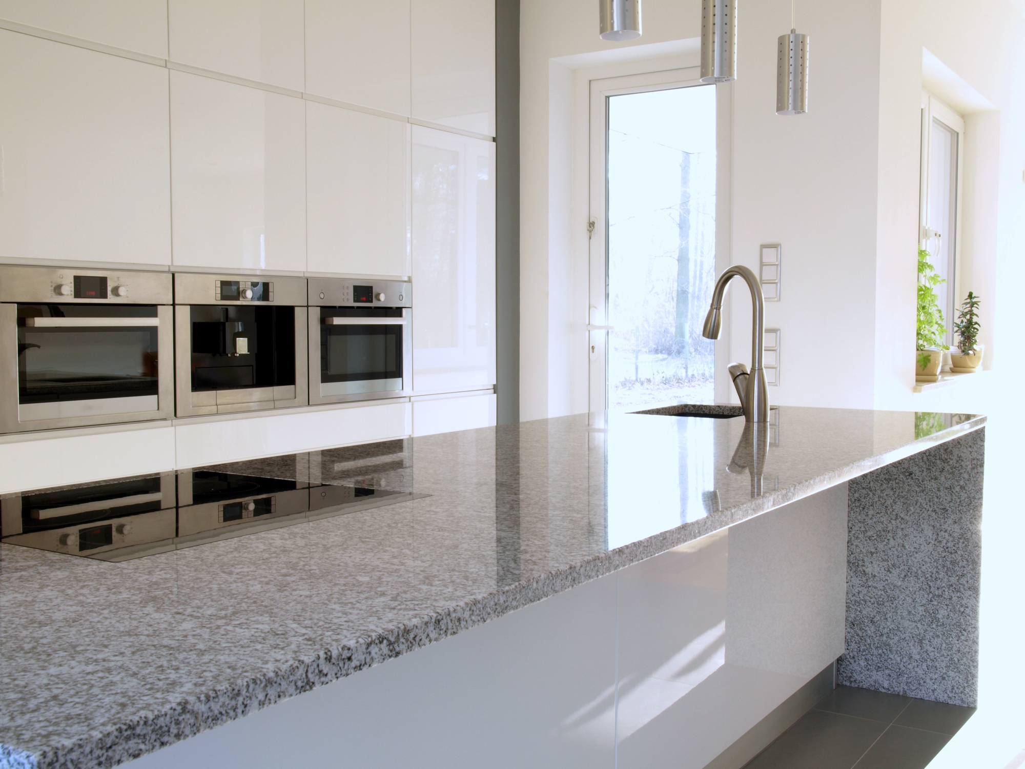 Modern kitchen with a polished granite island countertop, built-in ovens, an induction cooktop, a sink with a gooseneck faucet, and large windows letting in natural light.