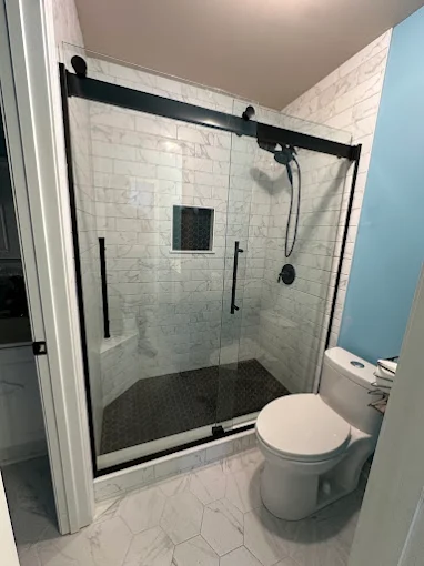 Modern bathroom with a glass-enclosed shower, white tile walls, black fixtures, and a toilet next to a light blue wall.
