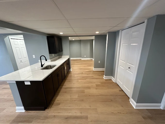 Modern basement kitchenette with dark cabinets, white countertops, light wood flooring, recessed lighting, and gray walls; white double doors are visible on the right.