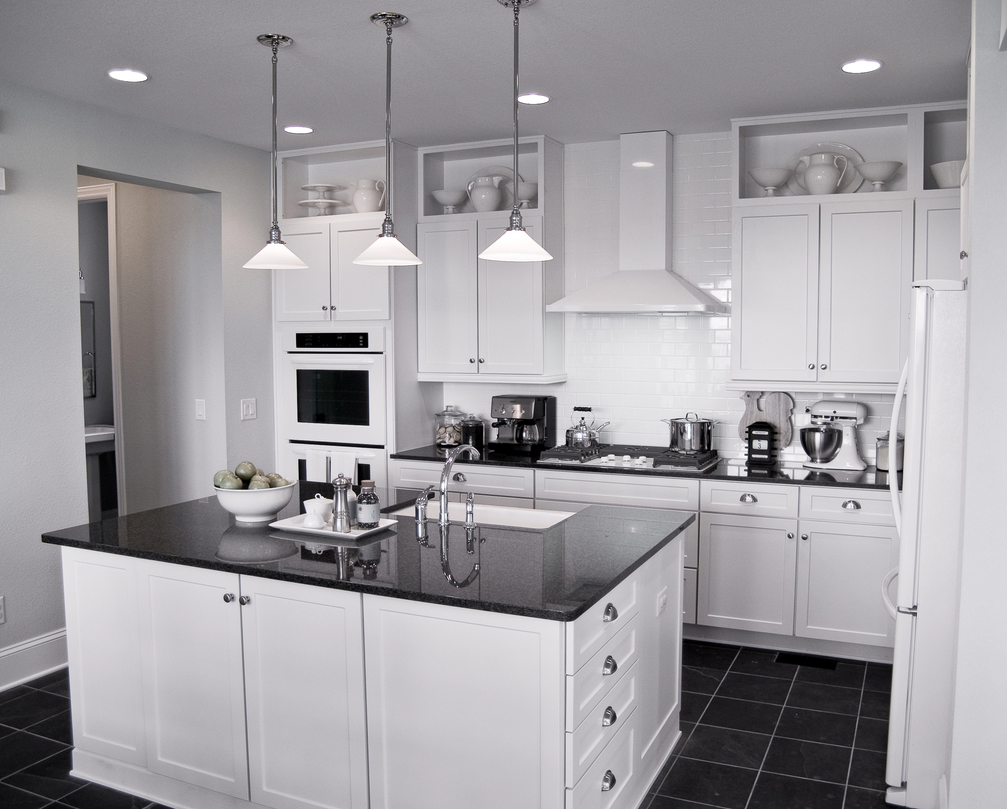 Modern kitchen with white cabinets, black countertops, a central island with a sink, pendant lights, and stainless steel appliances. Shelves hold white dishes and bowls.