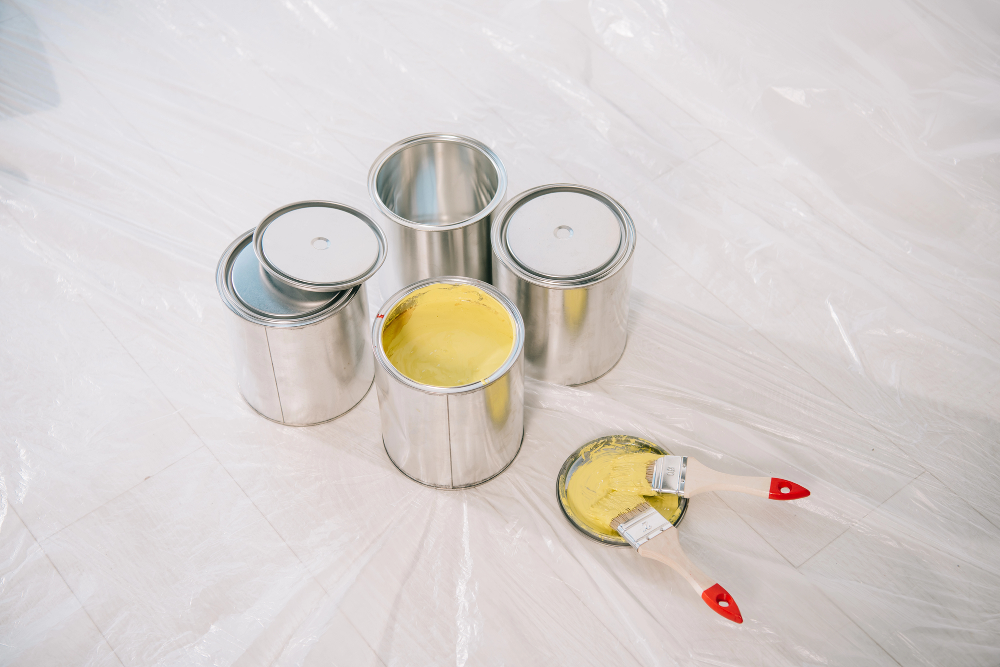 Four metal paint cans, one open with yellow paint inside, sit on a plastic sheet. Two paintbrushes with red handles rest on the open can’s lid covered in yellow paint.