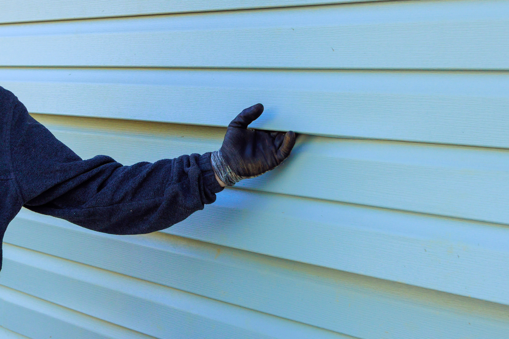 Person wearing a dark glove and sleeve touches light blue vinyl siding on a wall. Only the arm and hand are visible.